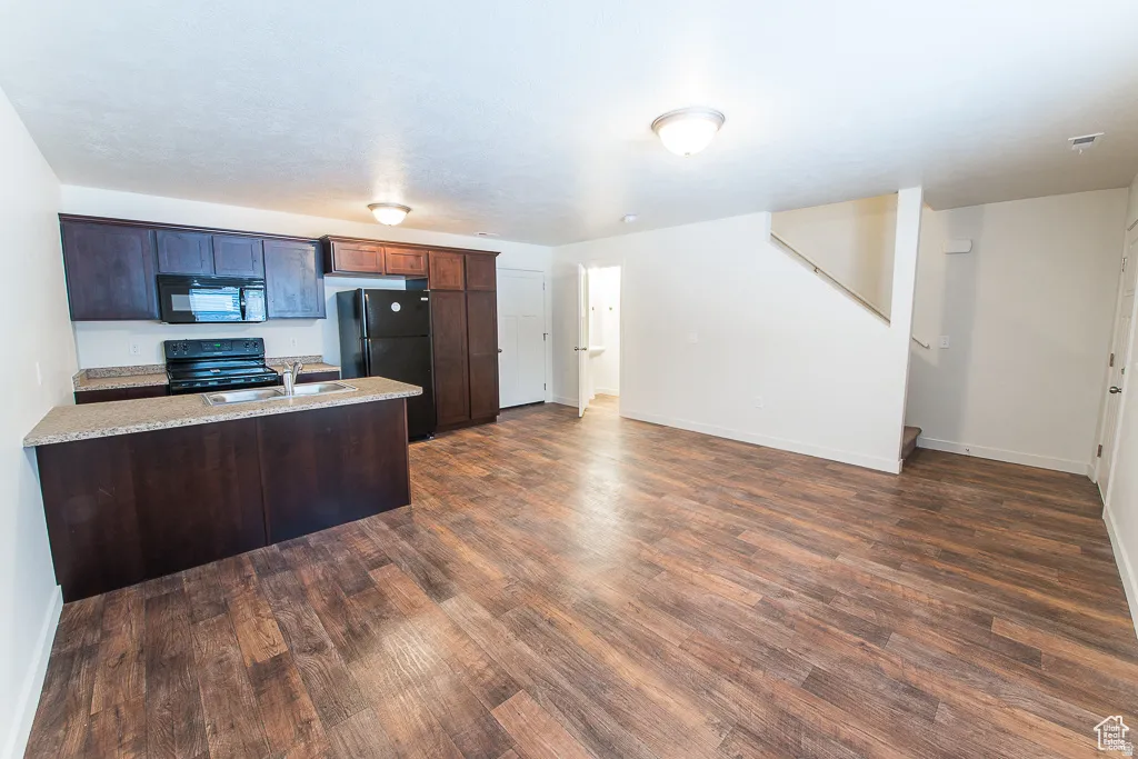 Open-concept living area featuring wood-finish flooring, dark wood cabinetry, black appliances, and a kitchen island with a double basin sink