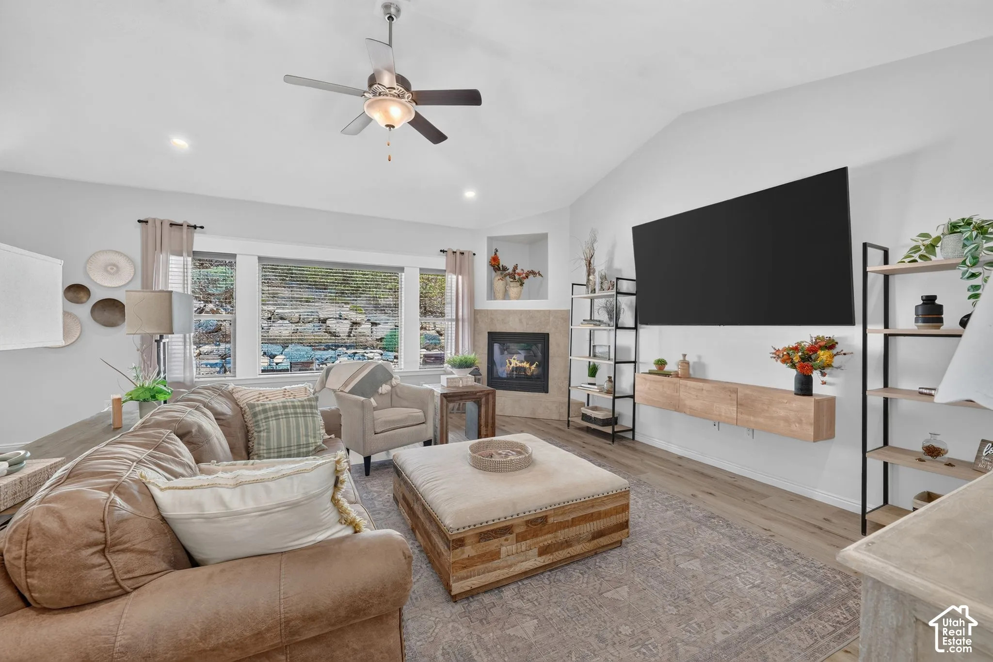 Vaulted ceiling with a ceiling fan, recessed lighting, fireplace with a stone surround, wood-finish flooring, and multiple windows