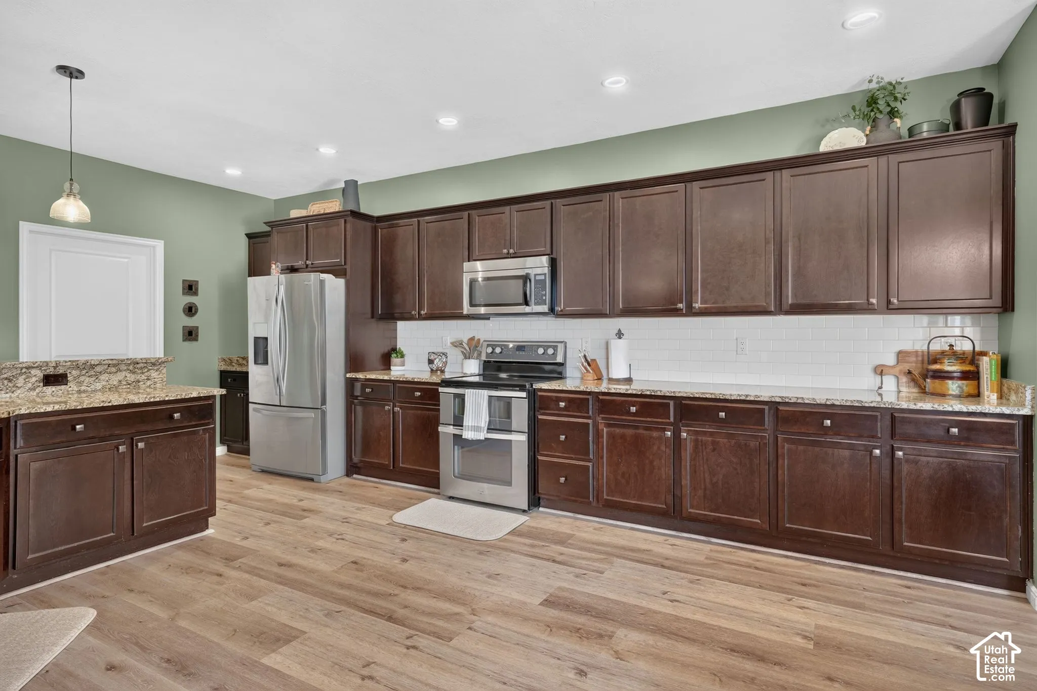 Spacious kitchen featuring extensive dark wood-finish cabinetry, light-toned wood-finish flooring, stainless steel appliances, granite-look countertops, and a subway tile backsplash