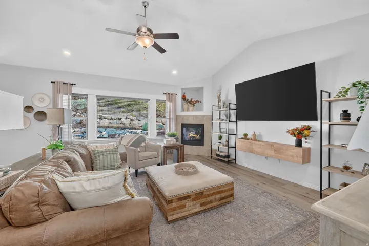 Vaulted ceiling with a ceiling fan, recessed lighting, fireplace with a stone surround, wood-finish flooring, and multiple windows
