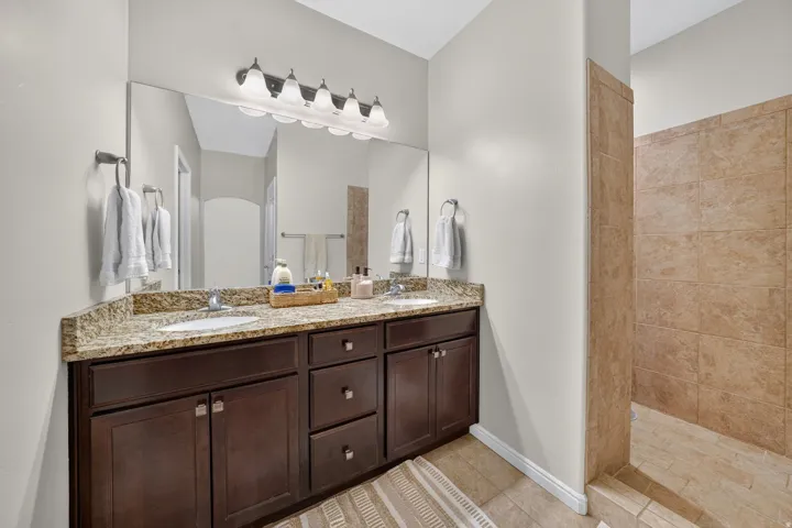 Double vanity with granite countertop, dual undermount sinks, wood-finish cabinetry, and a walk-in shower with tile surround