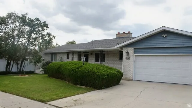 Brick exterior with blue siding accents, an attached garage, a paved driveway, a front lawn, and mature landscaping