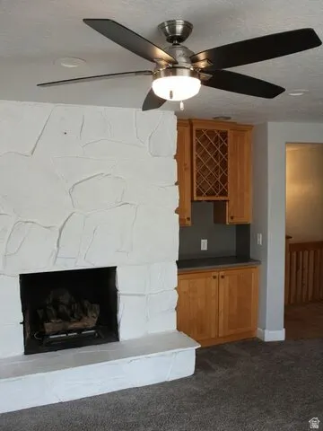Stone-clad fireplace with hearth, built-in cabinetry with a wine rack, ceiling fan with integrated lighting, and carpet flooring
