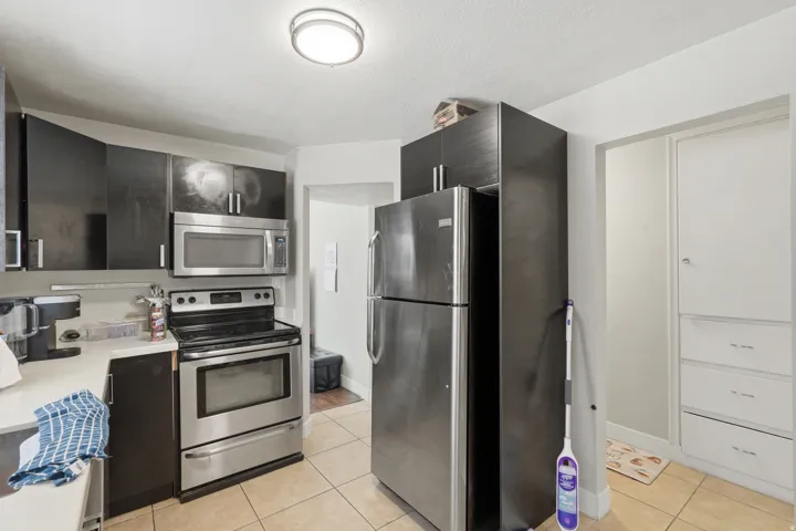 [Unit 2] Modern kitchen equipped with dark contemporary cabinetry, stainless steel appliances, neutral tiled flooring, and nearby built-in hall storage.