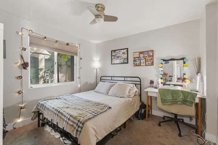 [Unit 1] Cozy bedroom featuring soft carpeting, an updated ceiling fan, and a large window providing ample natural light. The space comfortably accommodates a bed and a study desk.
