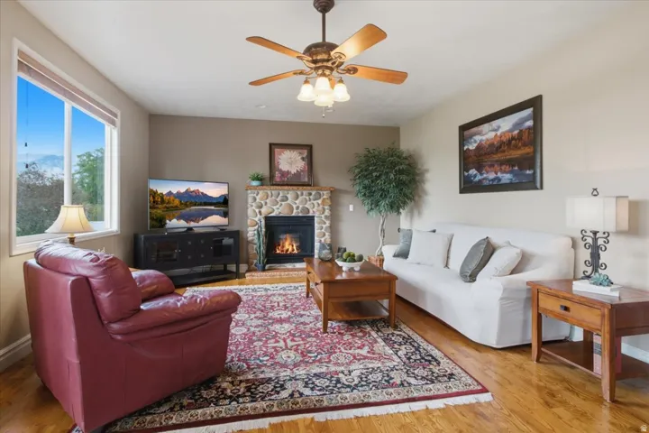 Wood-finish flooring, stone-clad fireplace, ceiling fan with integrated lighting, built-in window, and a built-in media console