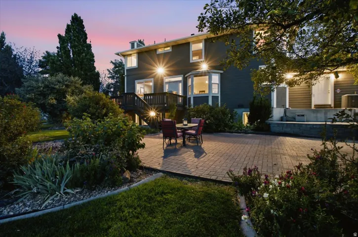 Expansive paver patio with a terraced retaining wall, surrounded by landscaped garden beds, a sloped lawn, and mature trees
