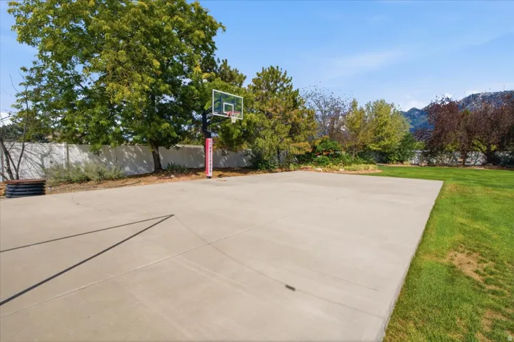 Expansive concrete sports court featuring a basketball hoop, surrounded by mature trees, a white perimeter fence, and a grassy lawn