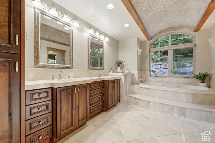 Dual vanity with stone countertops, wood-finish cabinetry, built-in soaking tub, arched window, and tile flooring