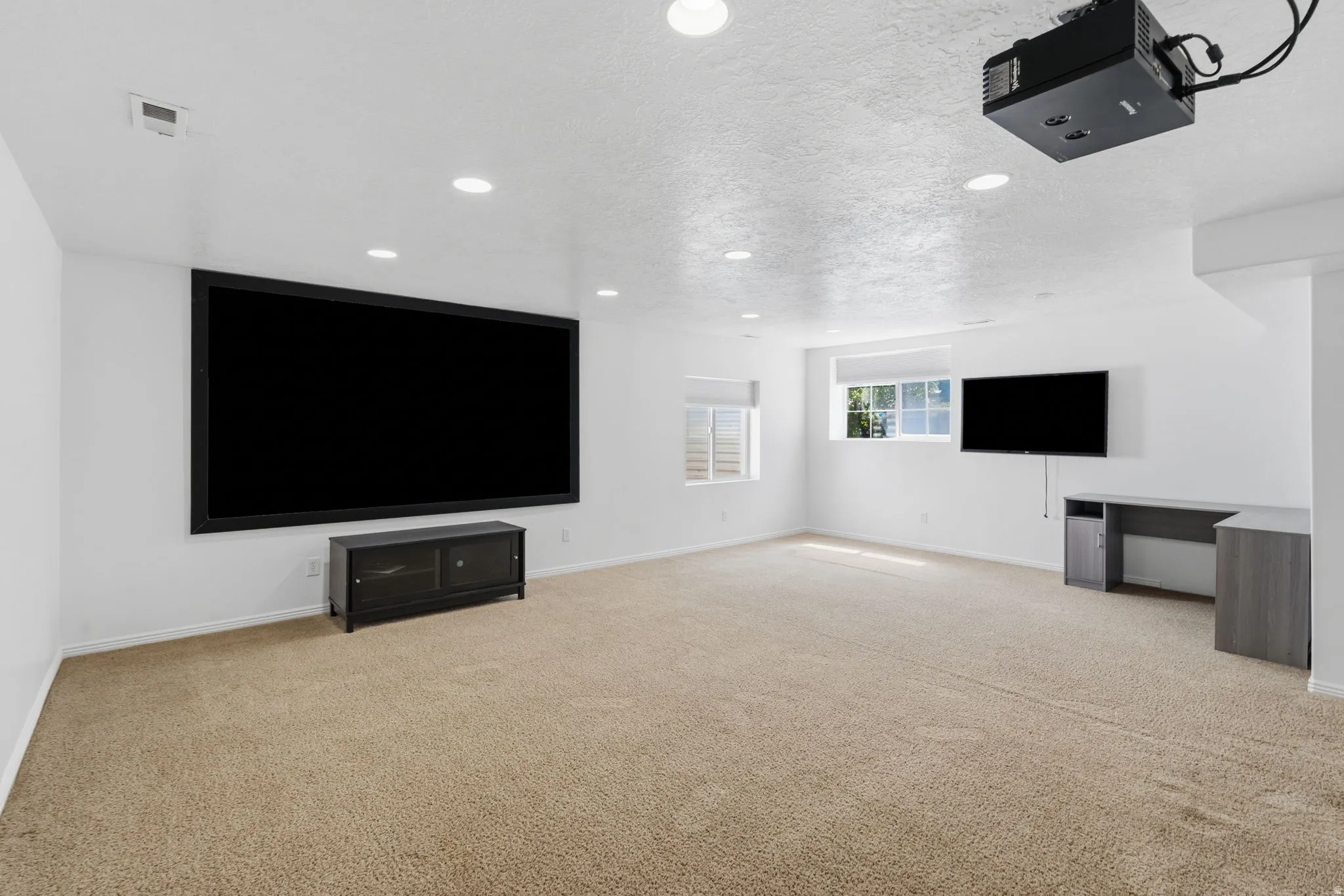 Recessed ceiling lighting, wall-mounted projector, large projection screen, carpet flooring, and multiple windows