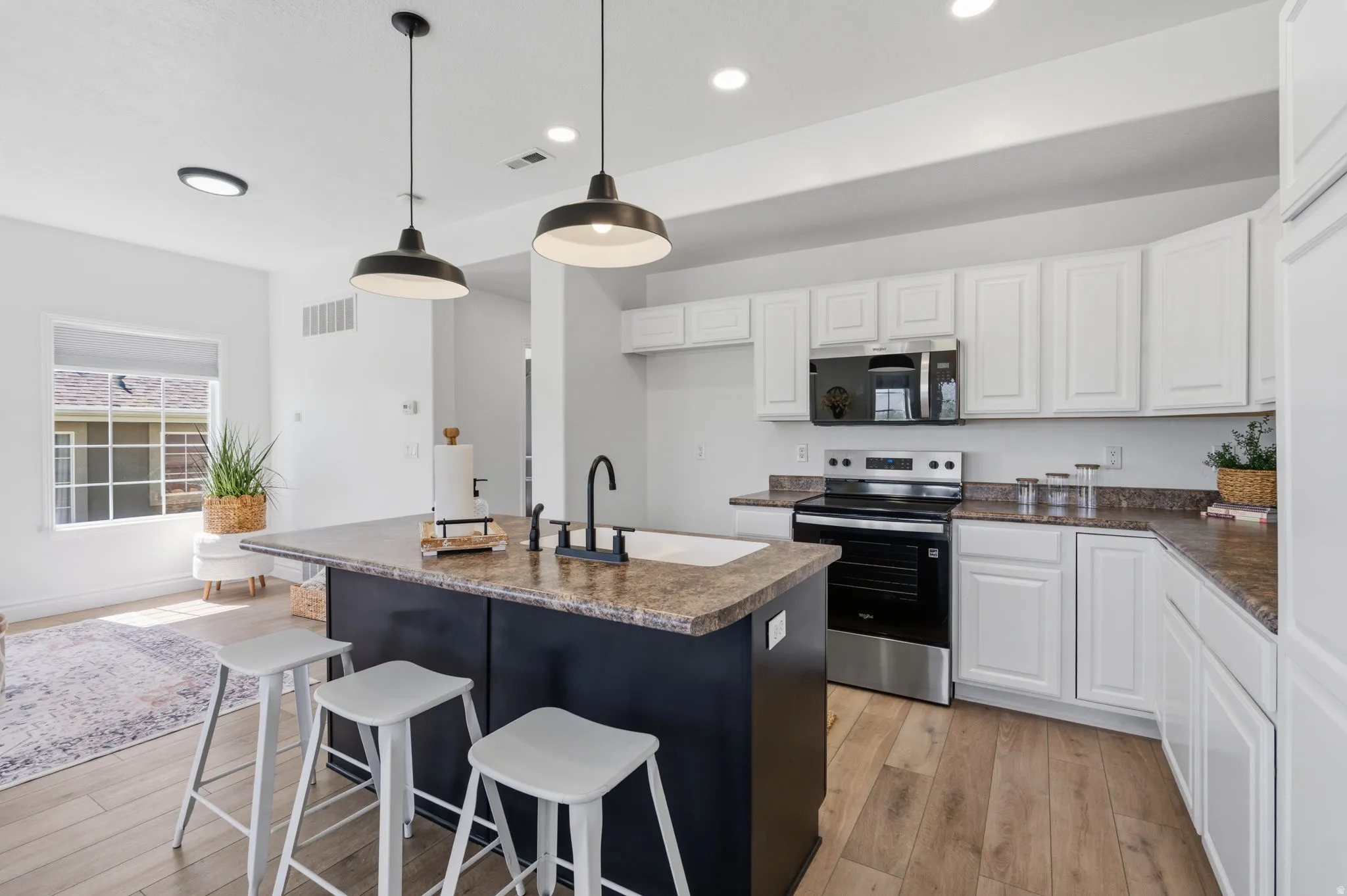 Kitchen featuring a dark-toned island with an integrated sink, white cabinetry, integrated stainless steel appliances, wood-finish flooring, and pendant lighting