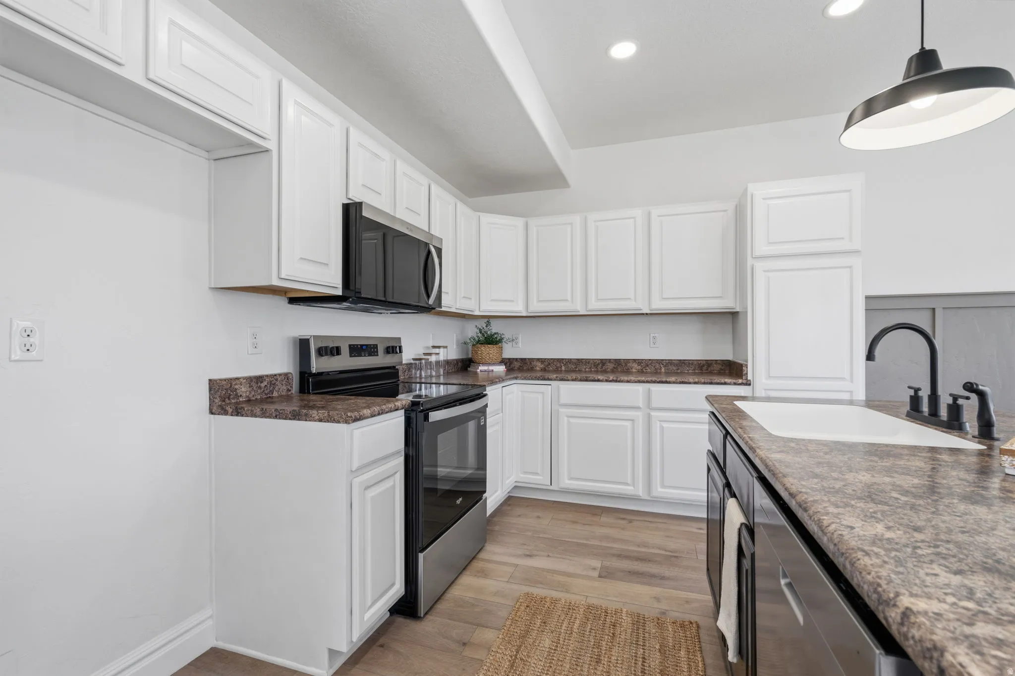White cabinetry with brushed nickel hardware, stainless steel appliances, wood-finish flooring, recessed lighting, and an island with a white sink and black faucet