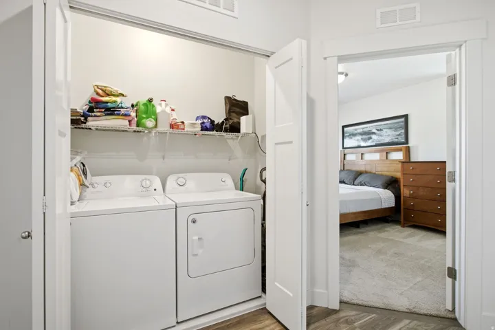 Laundry closet with built-in wire shelving, housing a white washer and dryer set, featuring wood-finish flooring