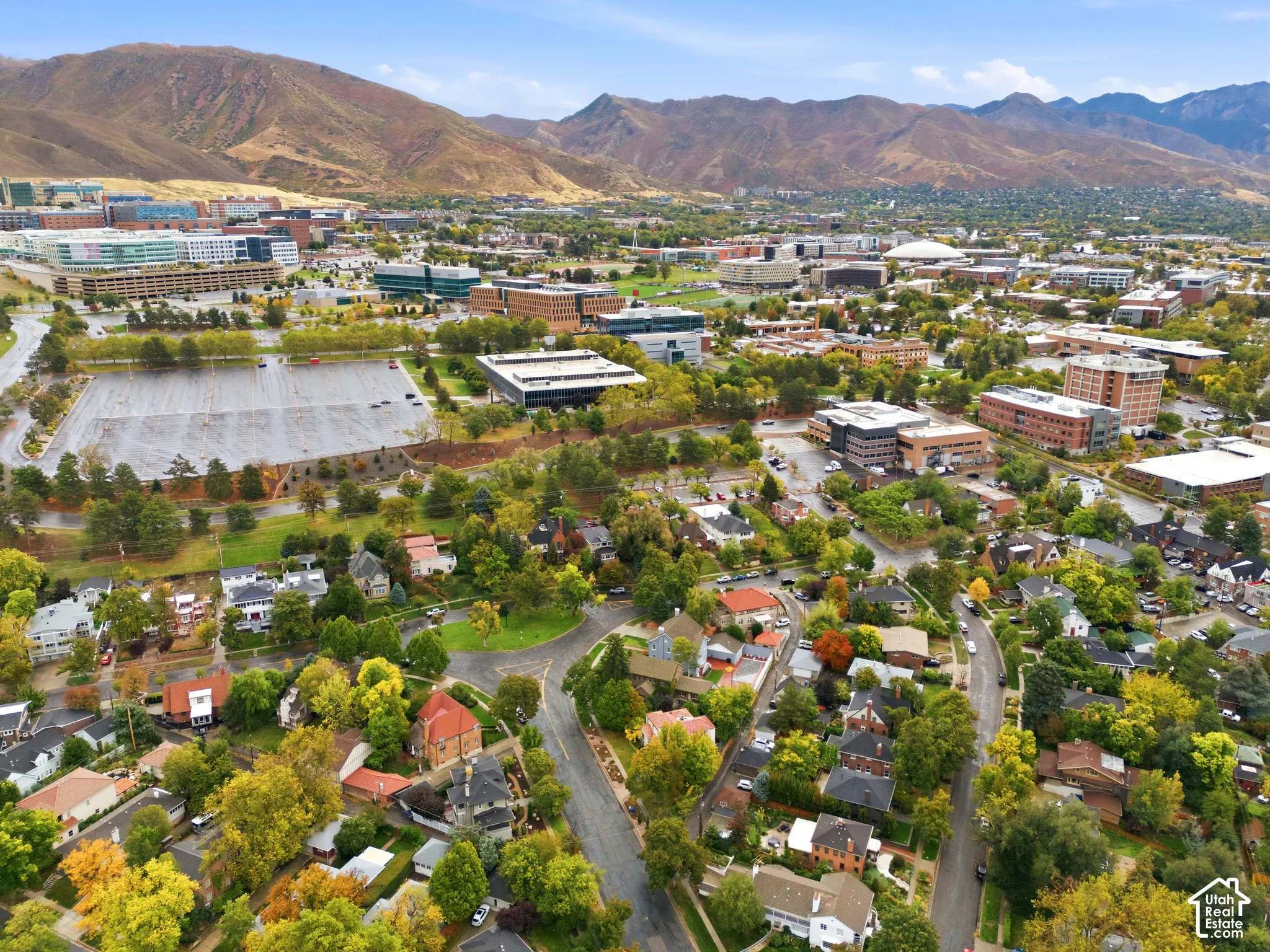 Expansive aerial perspective showcasing a residential area with mature trees, surrounded by commercial buildings and distant mountains under a blue sky