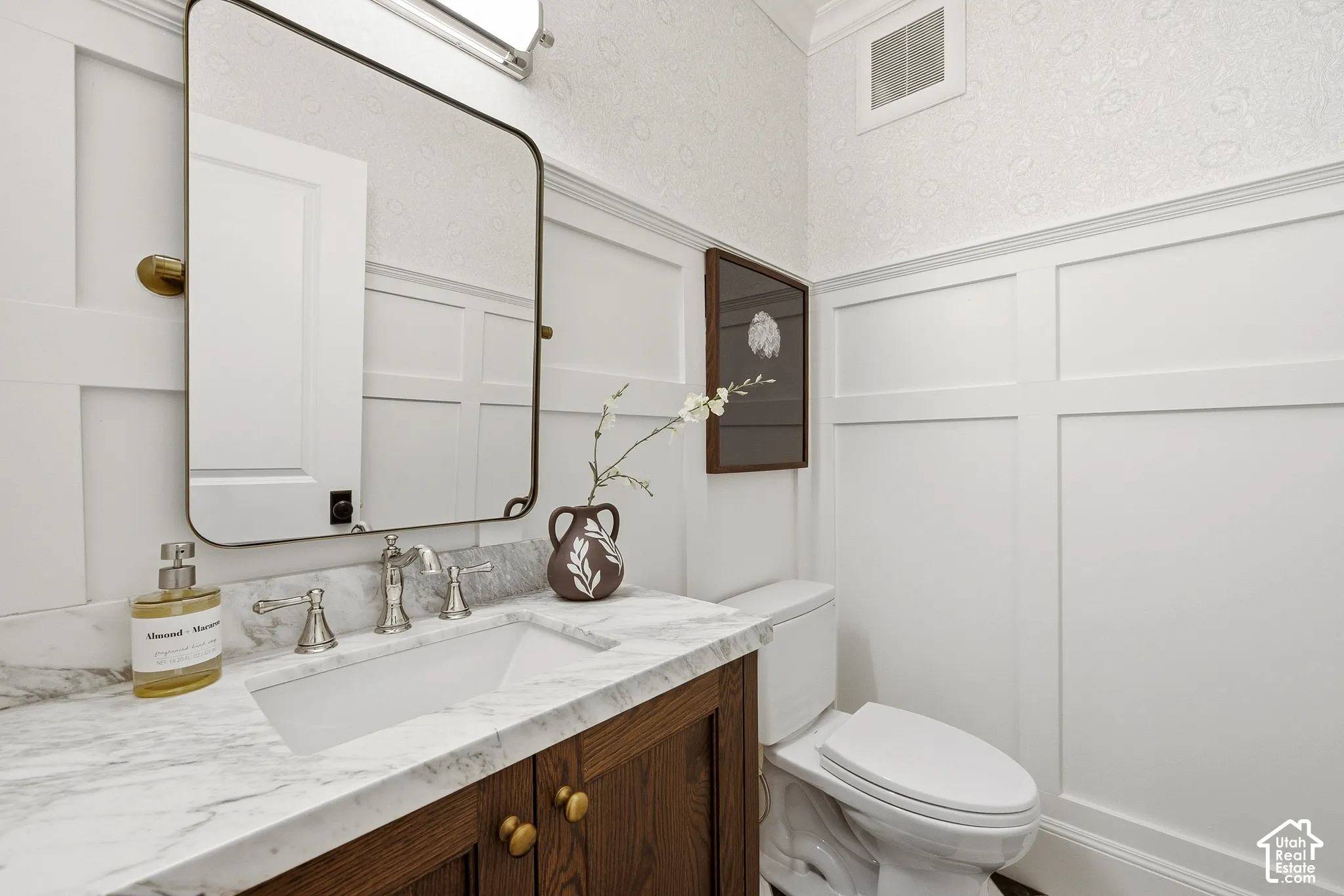 Bathroom featuring a wood-finish vanity with brass-toned hardware, an undermount sink, a stone countertop, waincotting, wallpaper, and polished nickel fixtures