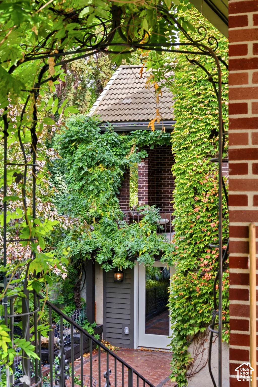 Wrought iron archway and railings over a brick paver patio, featuring a glass-paneled door, exterior wall lantern, and brick exterior with climbing vines