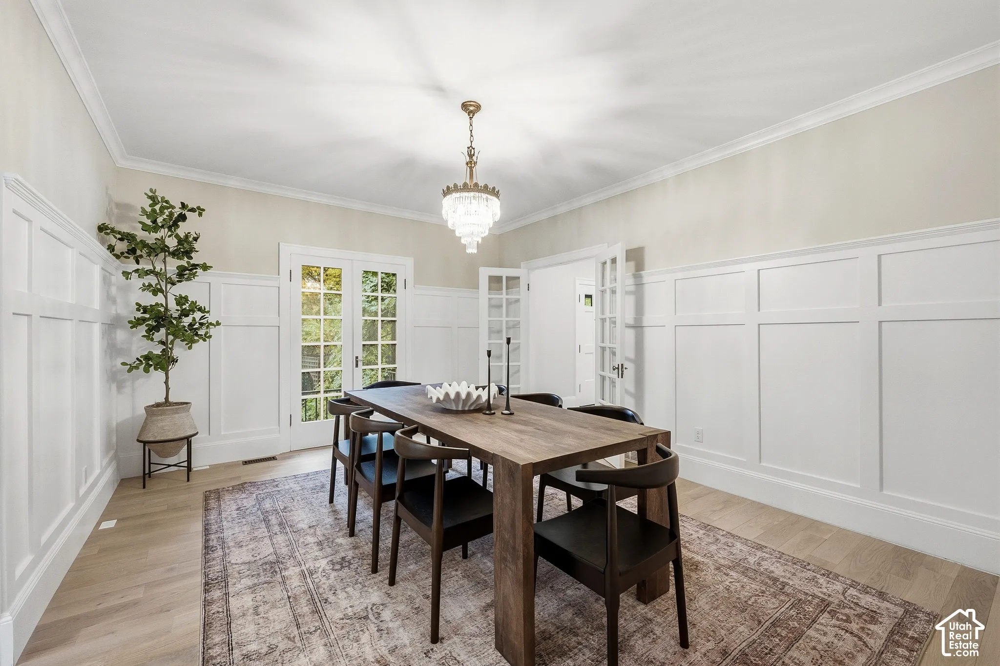 Crown molding, light wood-finish flooring, white wainscoting, French doors with multi-pane glass, and an antique crystal tiered chandelier