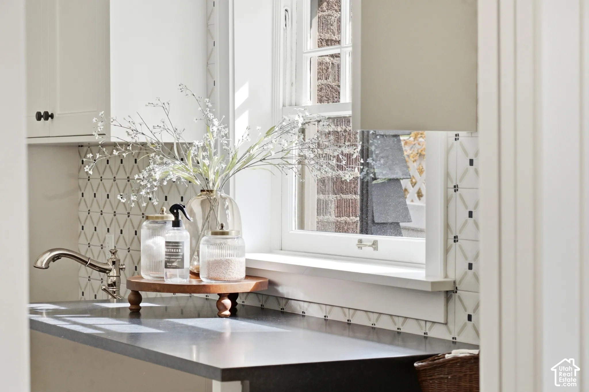 Creamy white cabinetry with bronze hardware, undermount sink with polished nickel faucet, patterned tile backsplash, black mist countertops, and double-hung window
