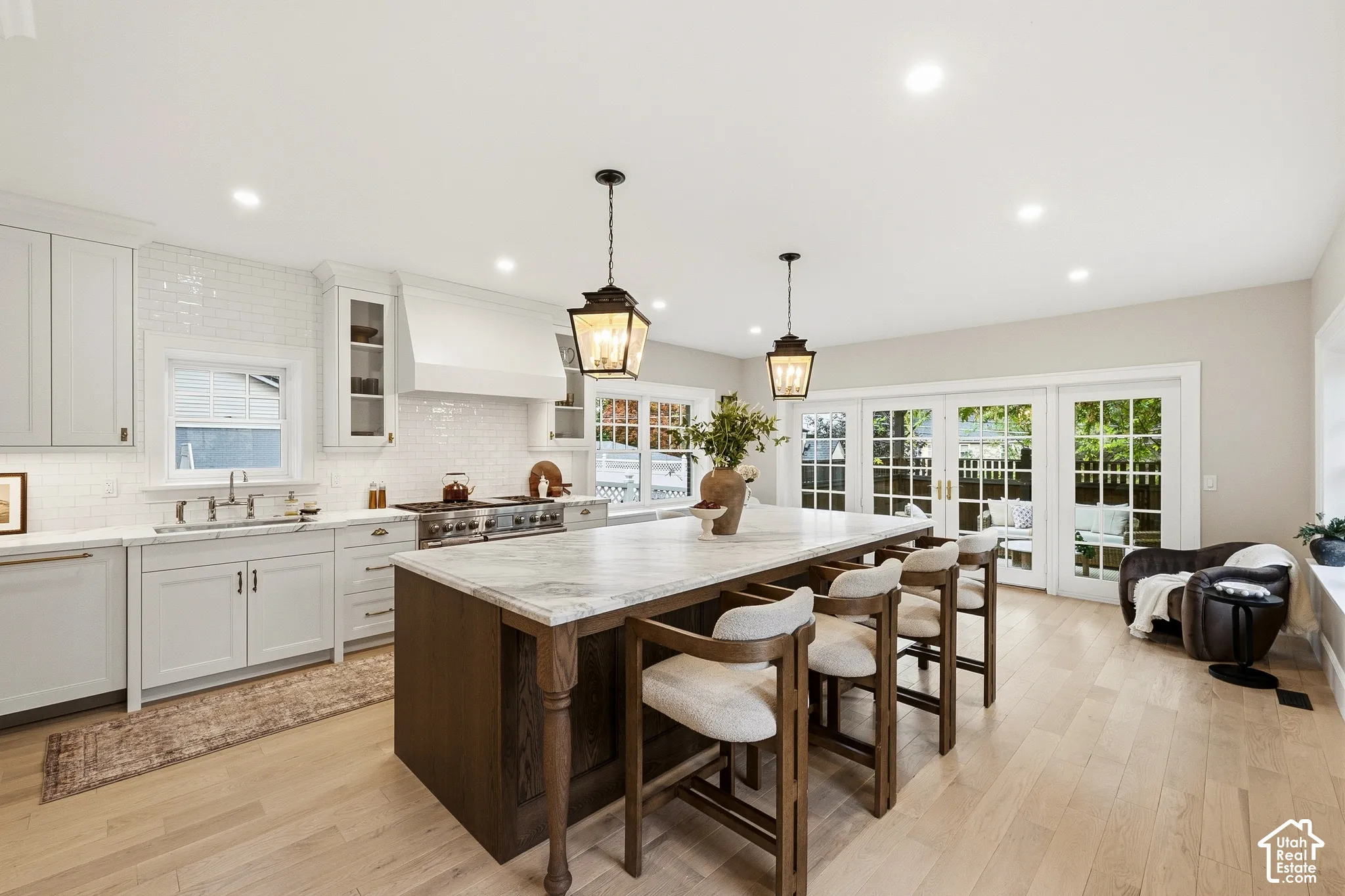 Spacious kitchen featuring light wood-finish flooring, a substantial kitchen island with a light-toned countertop, white cabinetry with traditional brass pulls, a white tile backsplash, and recessed lighting