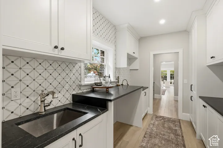 White shaker cabinetry, black countertops, undermount sink with gooseneck faucet, patterned tile backsplash, and light wood-finish flooring