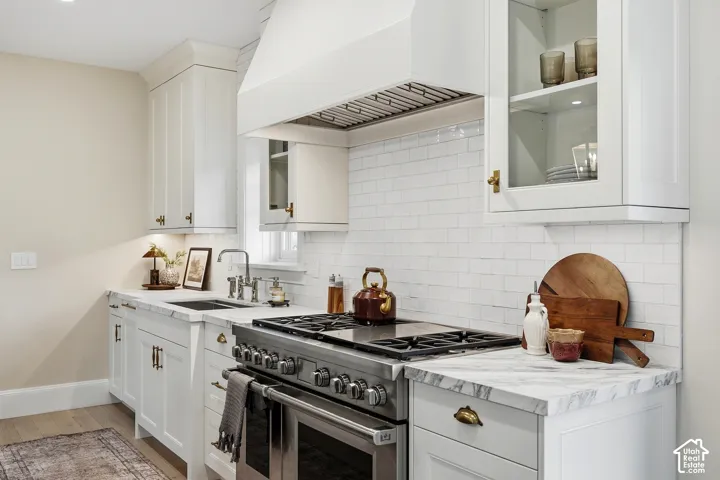 White shaker-style cabinetry with brass hardware, honed marble countertops, Wolf 48" stainless steel dual fuel range with a ventilation hood, subway tile backsplash, and a stainless steel sink with a polished nickel faucet