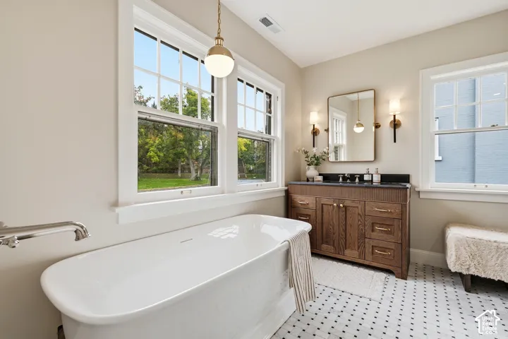 Primary Bath. Freestanding soaking tub, wood-finish vanity with dark countertop, multiple windows, patterned marble flooring, and wall-mounted sconces