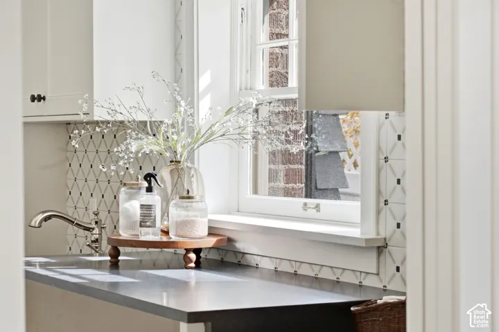 Creamy white cabinetry with bronze hardware, undermount sink with polished nickel faucet, patterned tile backsplash, black mist countertops, and double-hung window