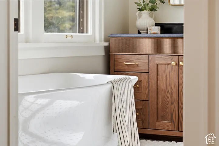 Freestanding soaking tub, wood-finish vanity with fluted detailing, solid surface countertop, gold-tone hardware, and casement window