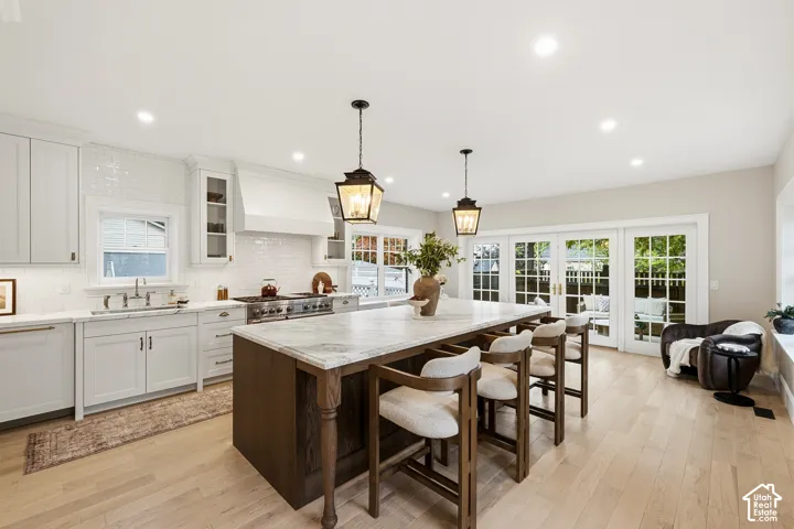 Spacious kitchen featuring light wood-finish flooring, a substantial kitchen island with a light-toned countertop, white cabinetry with traditional brass pulls, a white tile backsplash, and recessed lighting