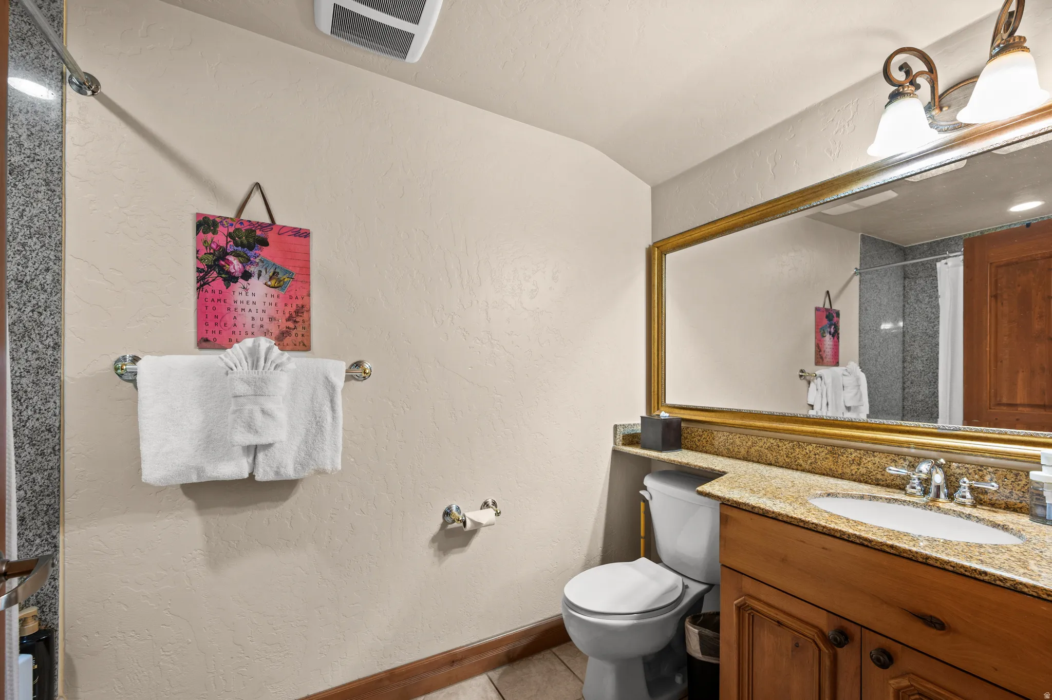 Elegant bathroom featuring warm wood cabinetry with ample storage, dual vanity with stone countertop and polished nickel fixtures, large framed mirror with brass trim, and tile flooring