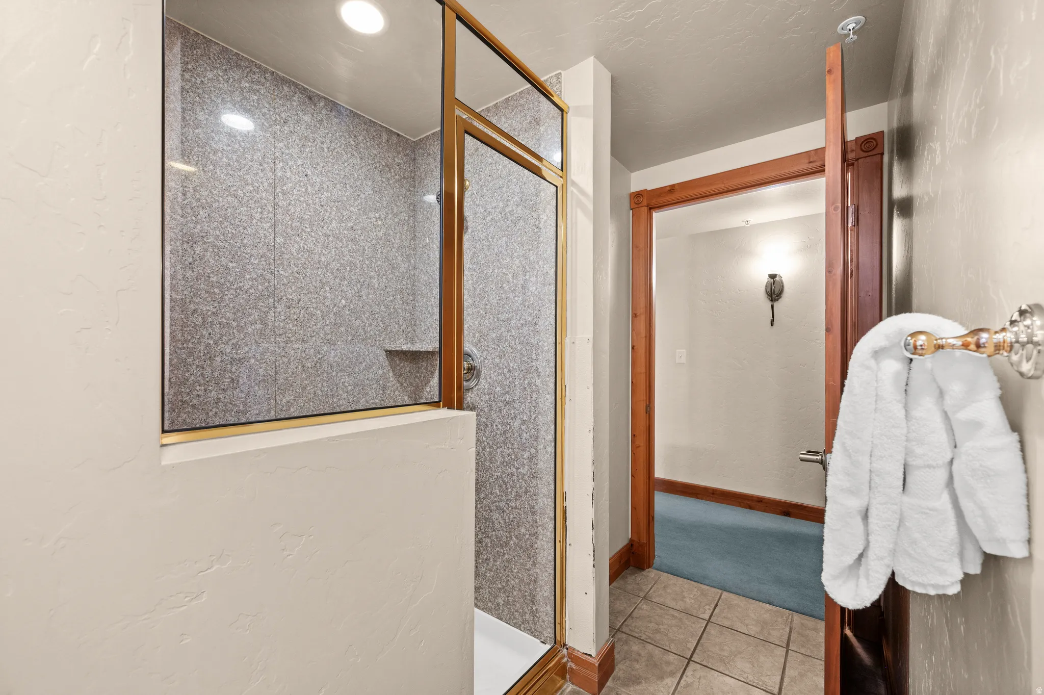 Primary Bathroom featuring a walk-in shower with textured tile surround and brass-finish framing, ceramic tile flooring, wood-finish door frame, and chrome towel bar with plush towels