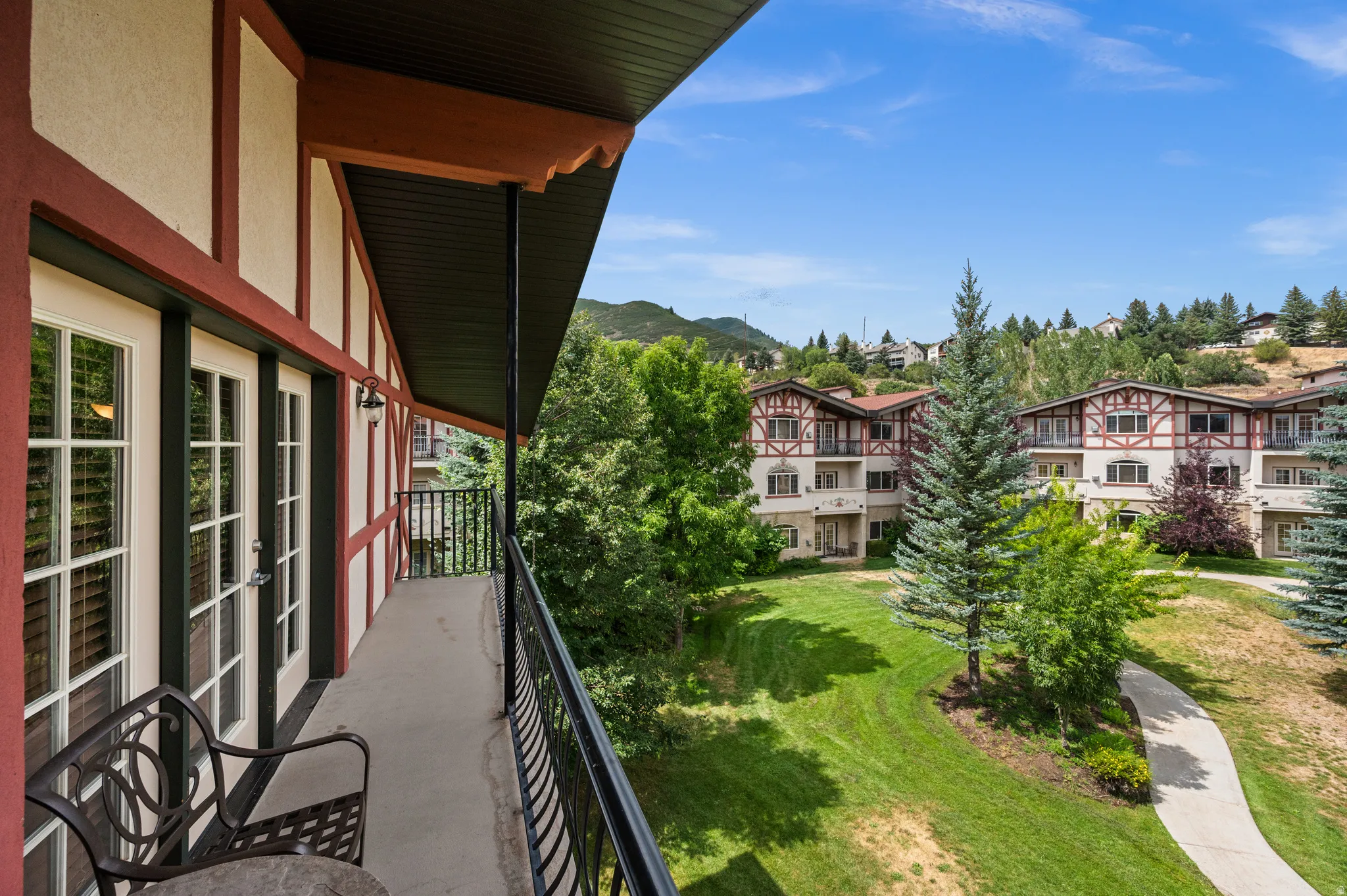 Expansive balcony with metal railing overlooking manicured grounds featuring mature evergreens and deciduous trees, rolling landscaping, and multi-unit residential architecture nestled against forested mountain backdrop