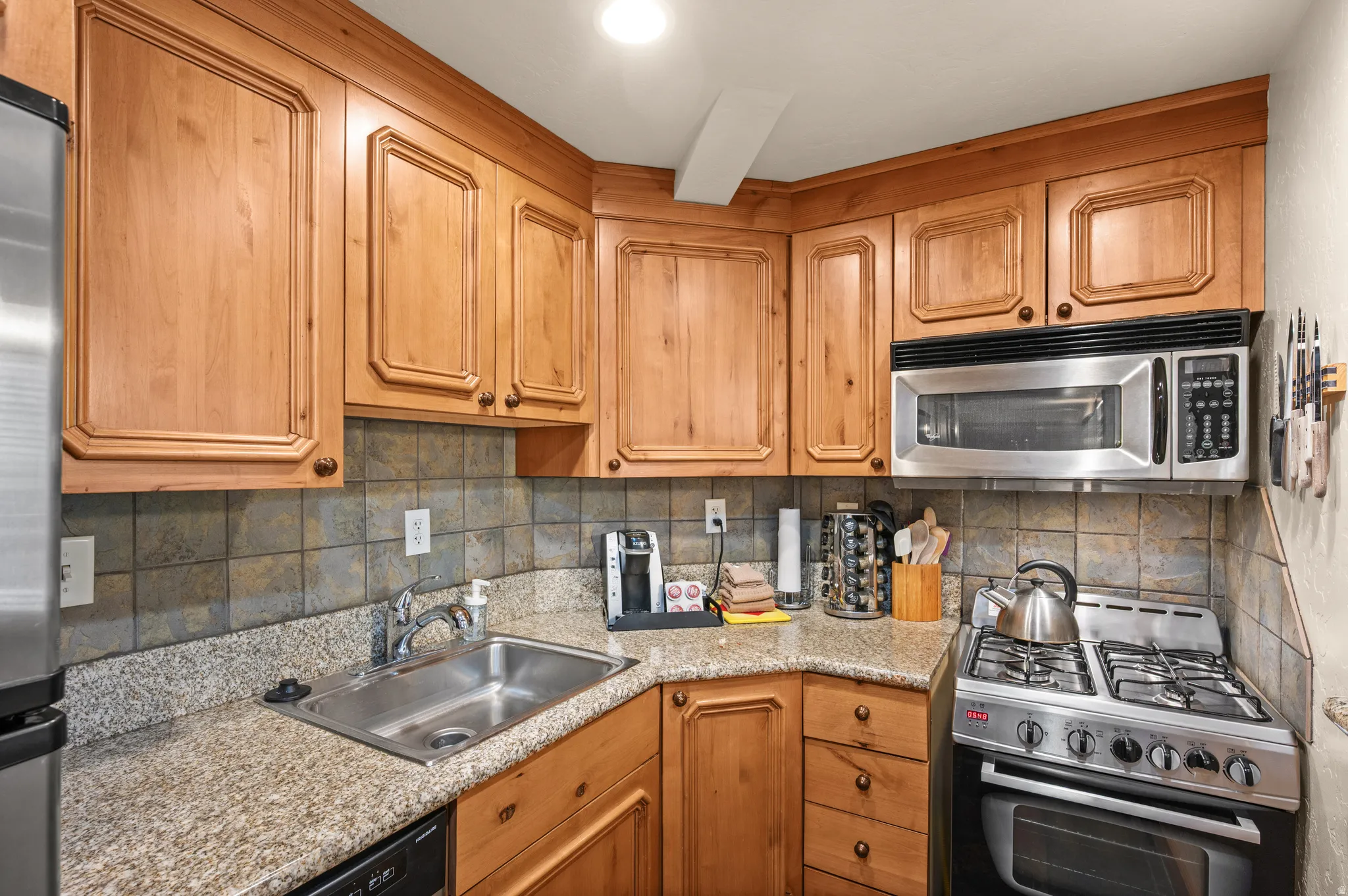 Wood-finish cabinetry, stainless steel sink, tile backsplash, granite-style countertops, and stainless steel appliances