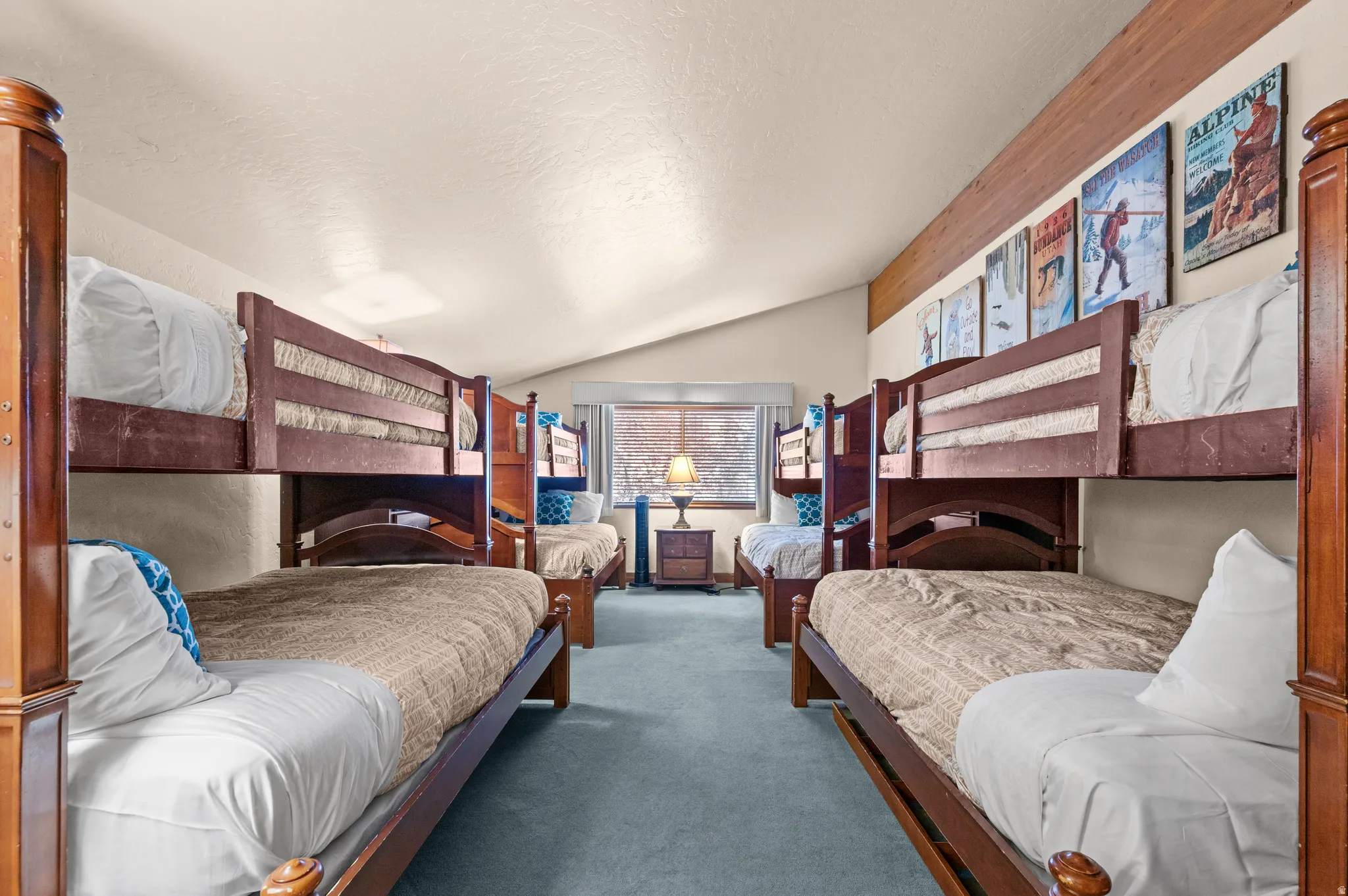 Spacious bunkroom featuring two wood-finish twin beds with burgundy headboards, blue-toned carpet flooring, recessed ceiling lighting, and decorative wall-mounted artwork