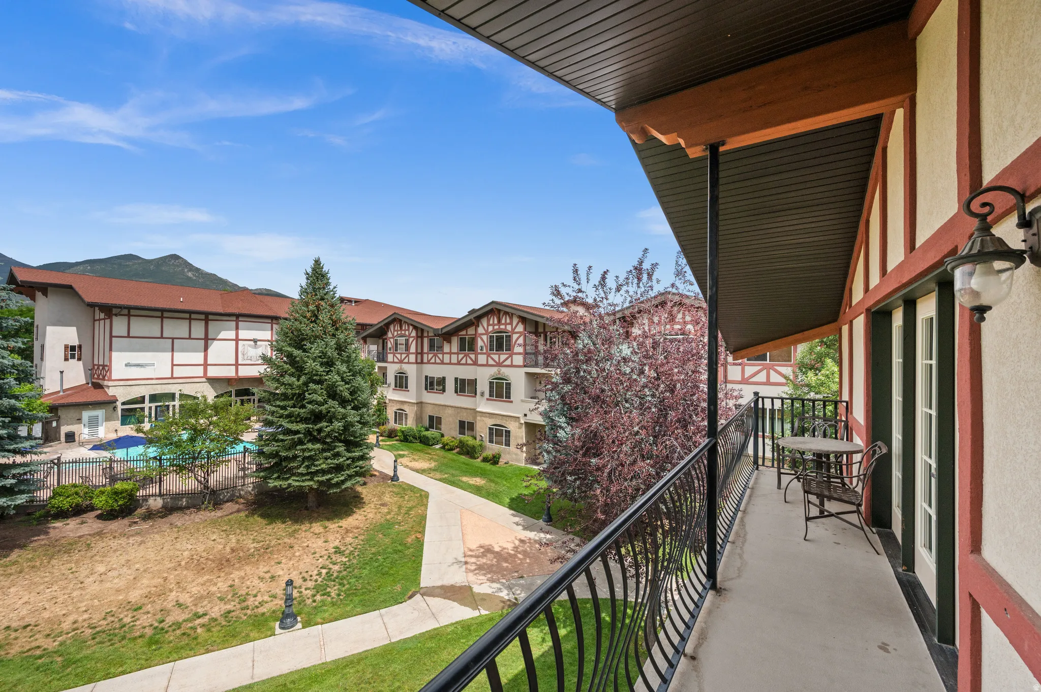 Elevated balcony with metal railings overlooking manicured grounds, mature evergreen landscaping, and resort-style amenities including a pool area