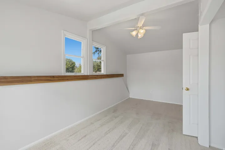 Sloped ceiling with exposed beams, ceiling fan with integrated lighting, two windows, wood-finish railing, and carpeted flooring