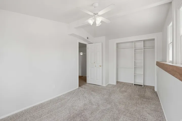Light-toned carpeting and white painted walls, ceiling fan with integrated lighting, built-in closet system with shelving and hanging rods, and wood-finish window ledge