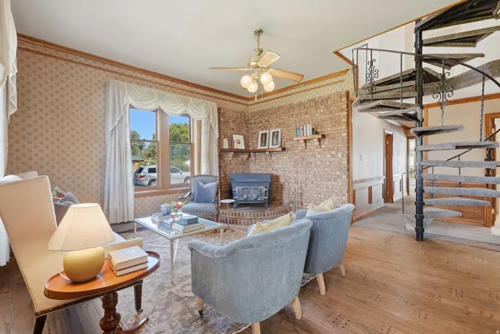 Wood-finish flooring, brick accent wall, wood-burning stove, spiral staircase with wrought iron railing, and a ceiling fan with integrated lighting