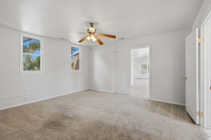 Neutral-toned carpeting, two windows with white trim, a brass-toned ceiling fan, and white walls