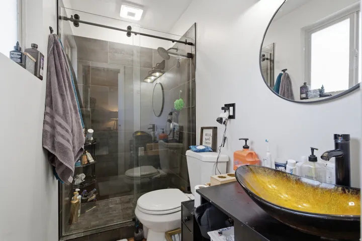 Modern bathroom featuring a glass-enclosed shower with dark-toned tile, a circular vanity mirror, and a vessel sink set on a dark wood-finish cabinet