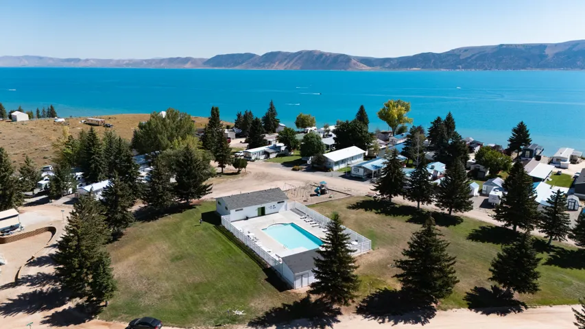 Community swimming pool with a white fence enclosure, surrounded by green lawn and mature trees, set against a Bear Lake and distant mountains.