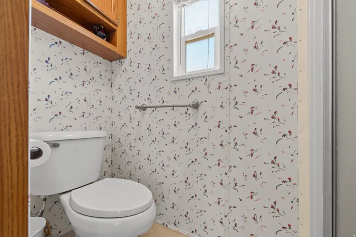 White toilet with a built-in toilet paper holder, wood-finish overhead storage, and walk in shower.