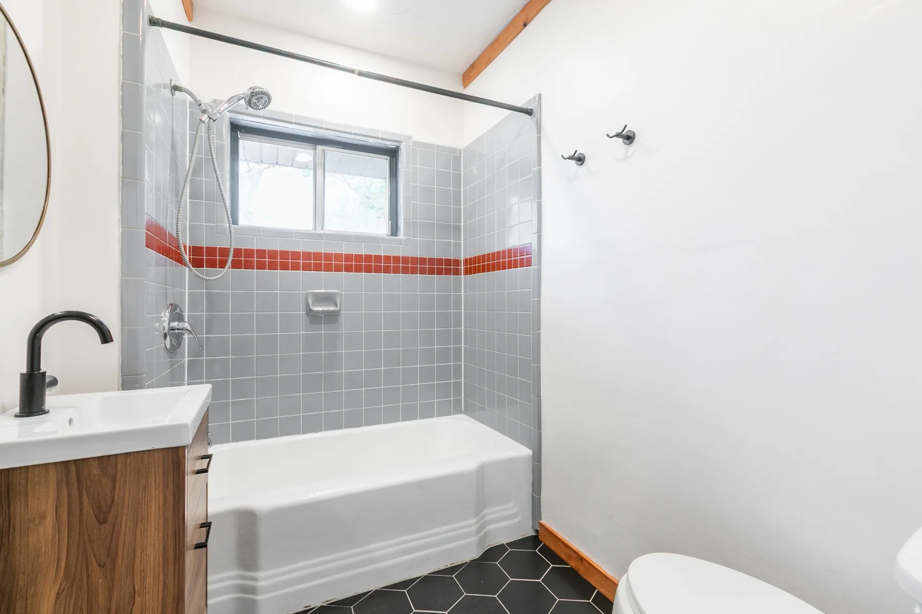 Bathtub with grey tiled surround and red accent stripe, wood-finish vanity with countertop sink and matte black faucet, and black hexagonal floor tiles
