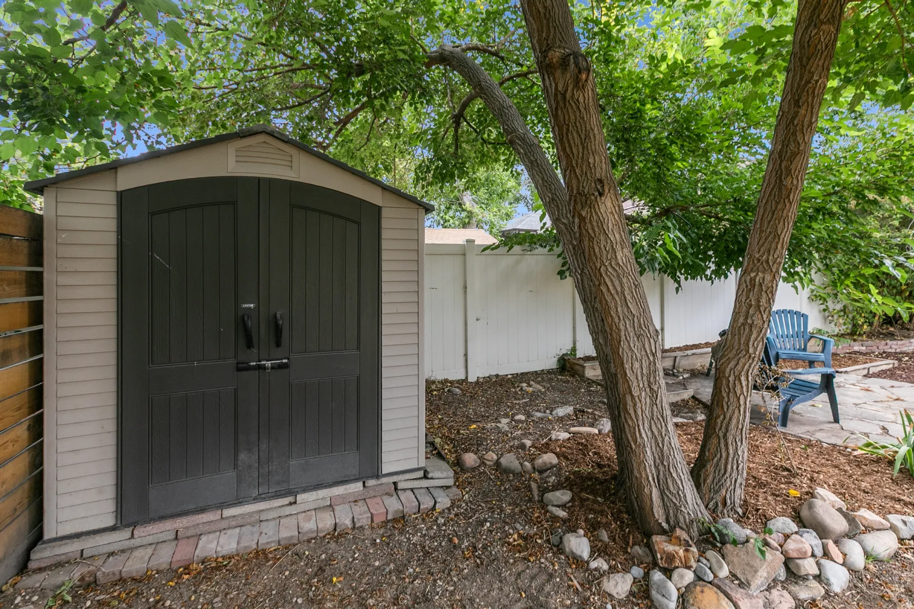 Storage shed featuring double doors, a white privacy fence, mature trees, and a flagstone patio area