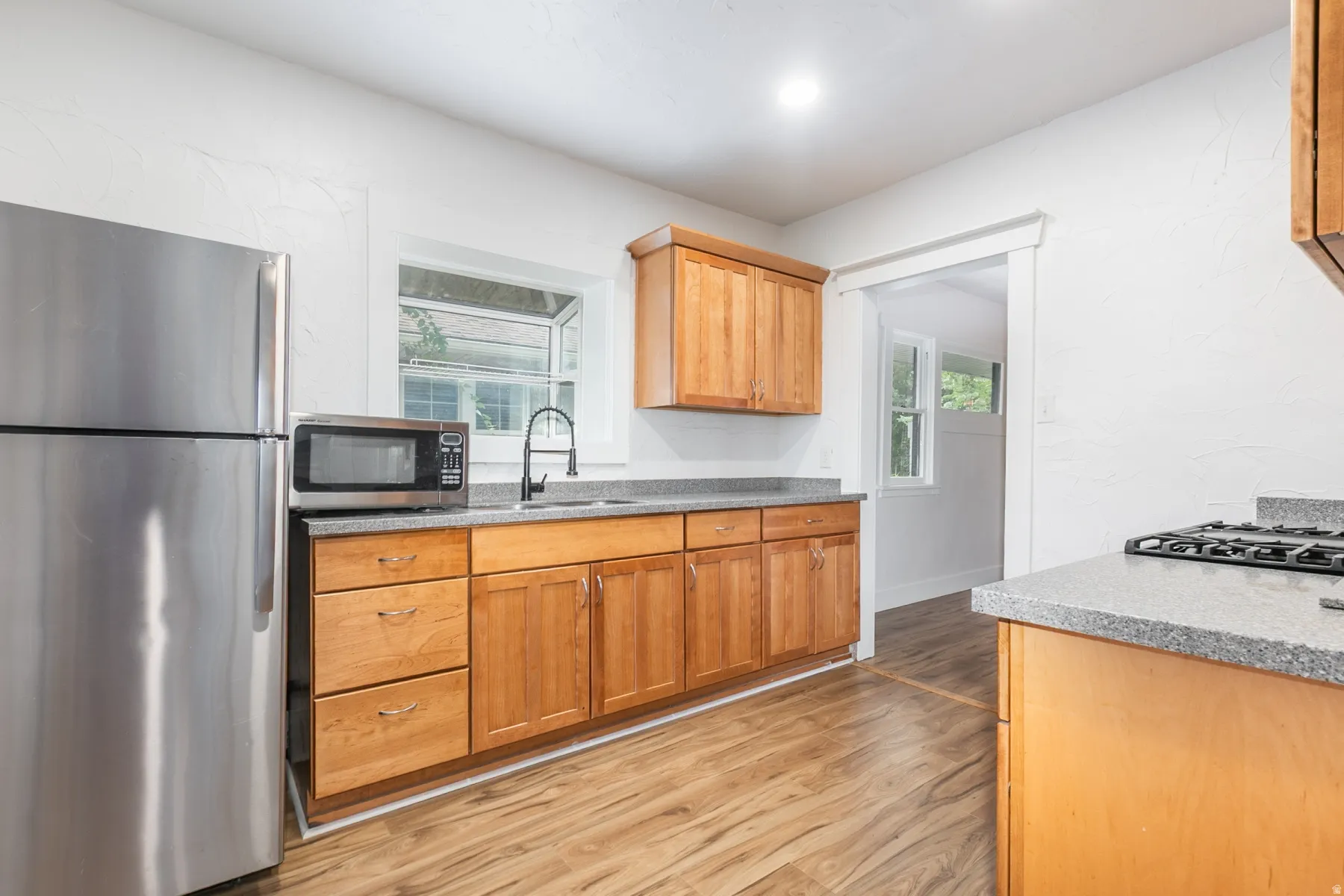 Stainless steel refrigerator, wood-finish flooring, light wood cabinetry, stone-look countertops, and stainless steel sink with black faucet
