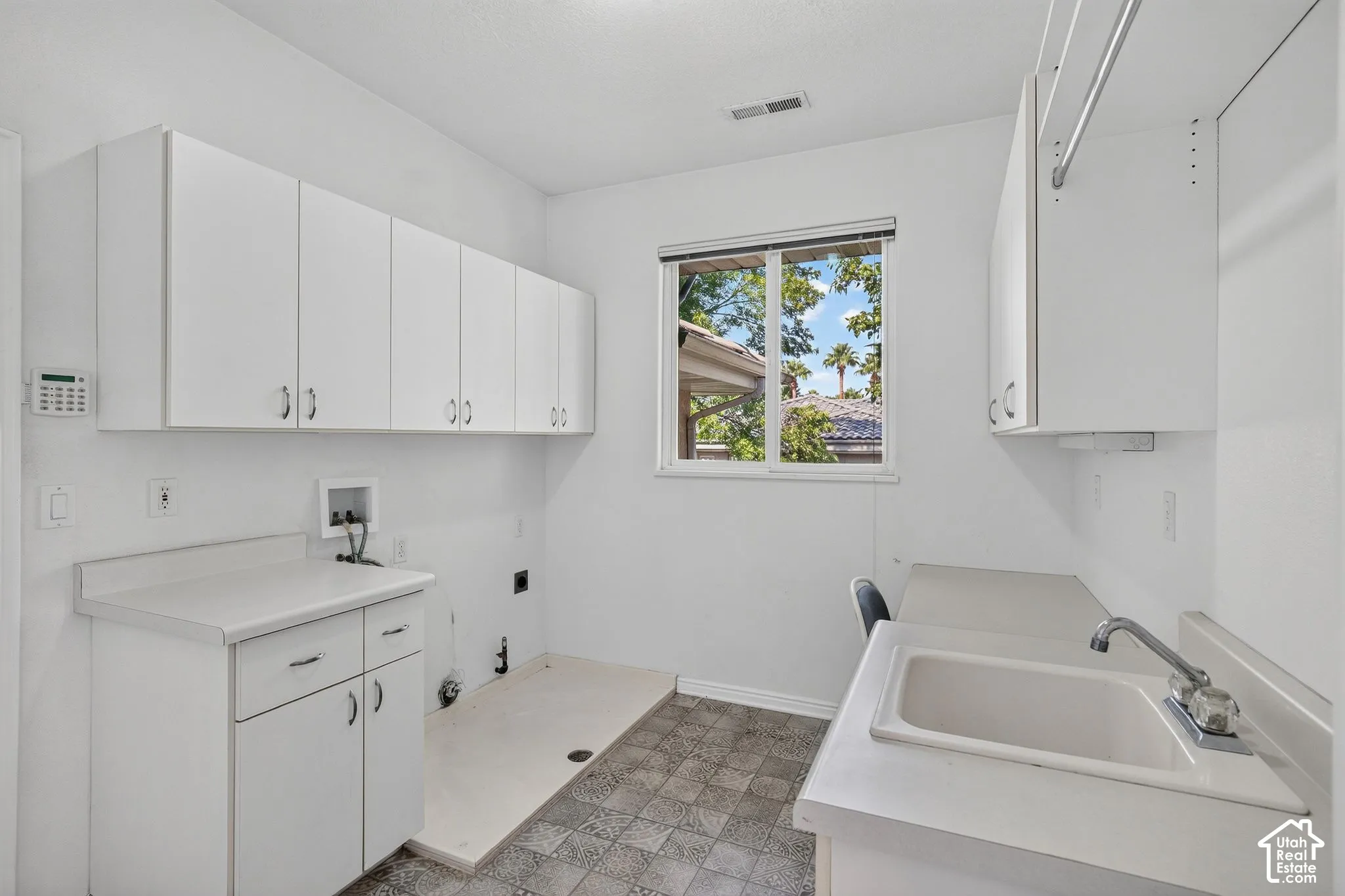 Bright laundry room featuring expansive white cabinetry with ample storage, dual stainless steel sink basins with chrome faucetry, patterned tile flooring, and abundant natural light from a large picture window overlooking outdoor landscaping