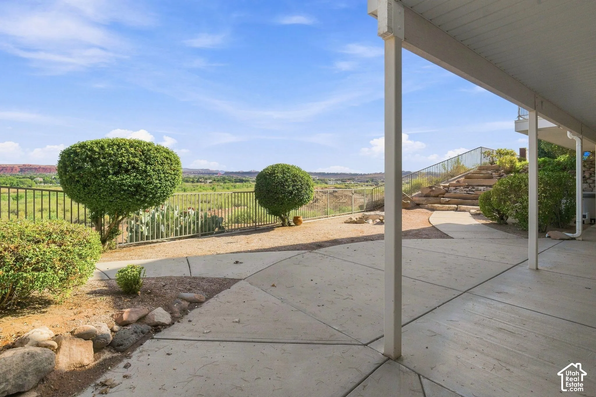 Expansive concrete patio with a covered outdoor area, terraced landscaping, metal fencing, and established shrubbery