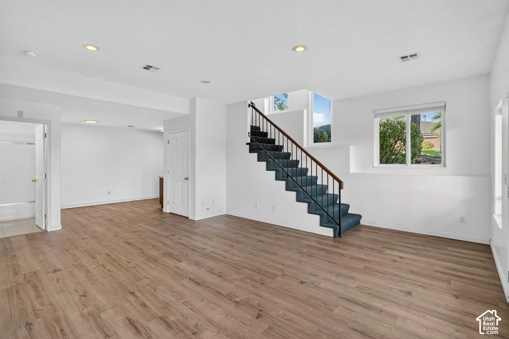 Expansive room featuring wood-finish flooring, recessed lighting, a carpeted staircase with an iron railing, and multiple windows