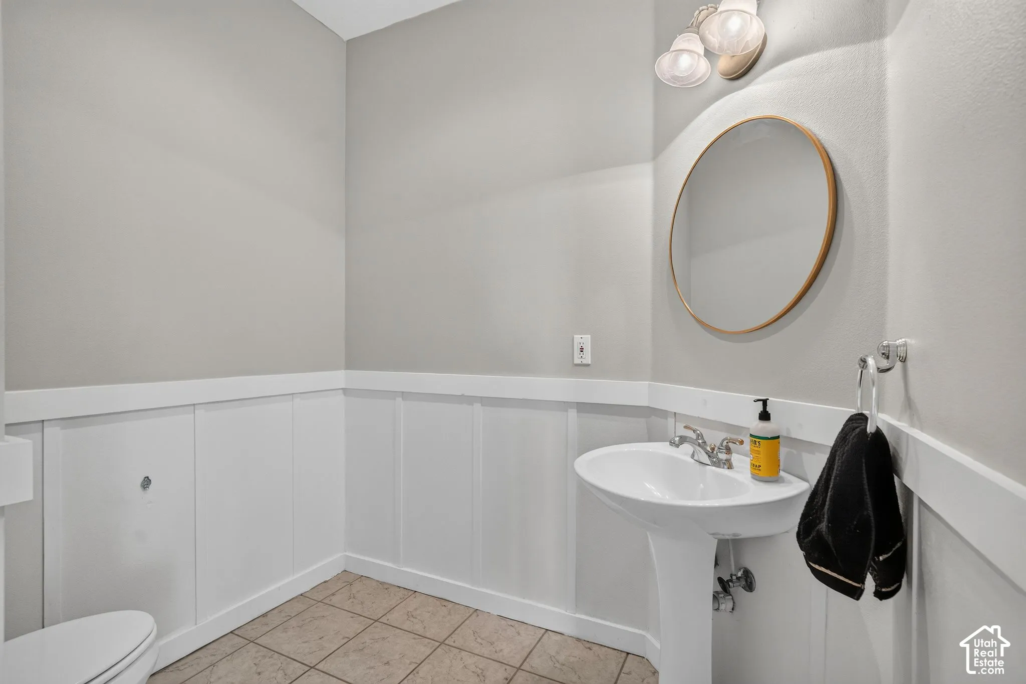 Pedestal sink with chrome faucet, circular mirror, wall-mounted light fixture, tile flooring, and white wainscoting