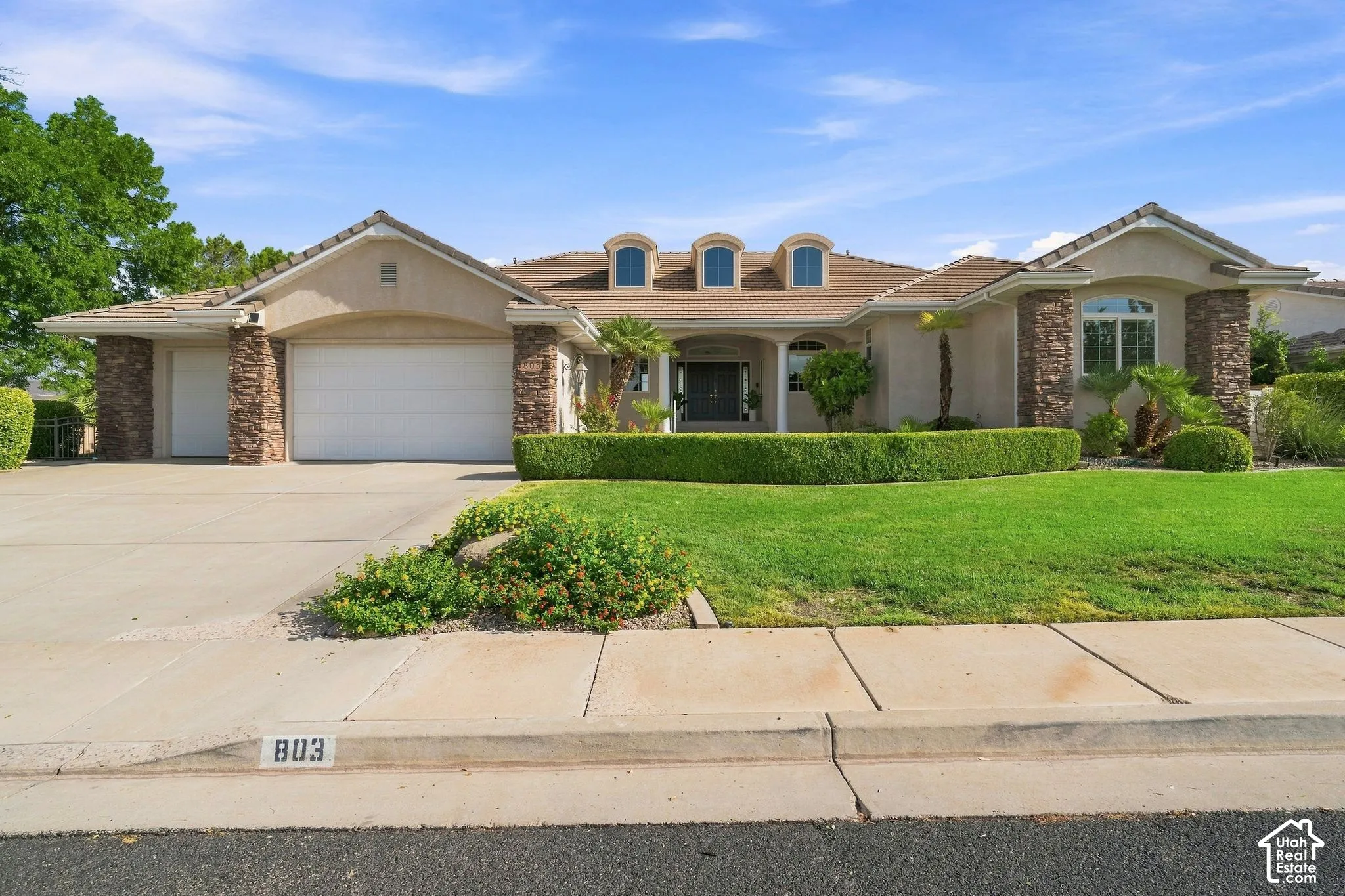 Elegant single-story residence featuring a stucco and brick exterior with multiple roof dormers, two-car garage, and manicured front landscaping with mature trees and hedging
